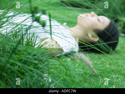 Donna sdraiata su erba, gli occhi chiusi, focus su piante in primo piano Foto Stock