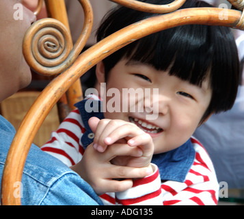 Sorridente, ragazzino, forse ragazza, sedette sulle ginocchia del padre. Visto attraverso le braccia di una sedia cinese di bentwood. Fuori dal Campanile Hotel, Xian. Foto Stock