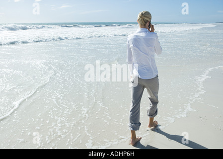 A piedi nudi imprenditrice a piedi attraverso il surf sulla spiaggia, utilizzando il telefono cellulare Foto Stock