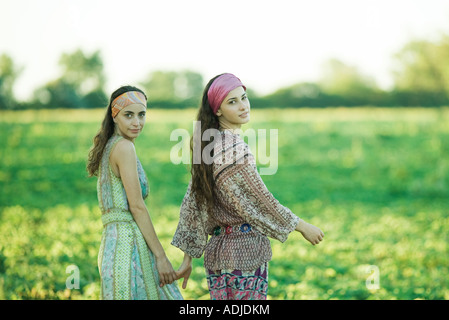 Giovani donne hippie camminare attraverso il campo, tenendo le mani, guardando sopra le spalle alla telecamera Foto Stock