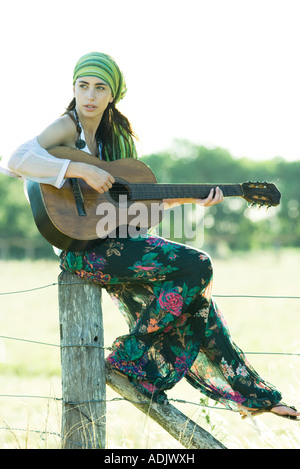 Giovani hippie donna seduta sul palo da recinzione, suonare la chitarra Foto Stock