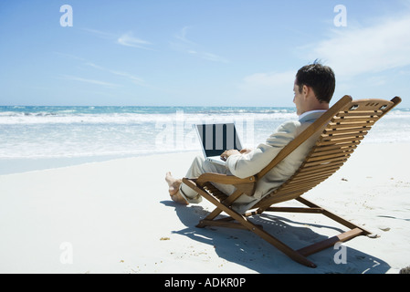 Imprenditore seduto nella sedia a sdraio in spiaggia a piedi nudi, utilizzando laptop Foto Stock