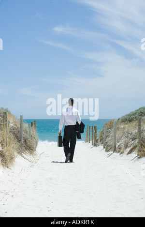 Imprenditore passeggiate verso la spiaggia, valigetta di contenimento e camicia al di sopra del braccio, vista posteriore Foto Stock