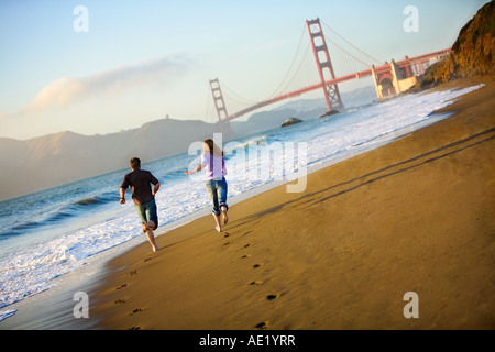 Un paio di correre sulla spiaggia verso il Golden Gate bridge lasciando tracce sulla spiaggia di sabbia dorata e delle onde oceaniche in crash Foto Stock