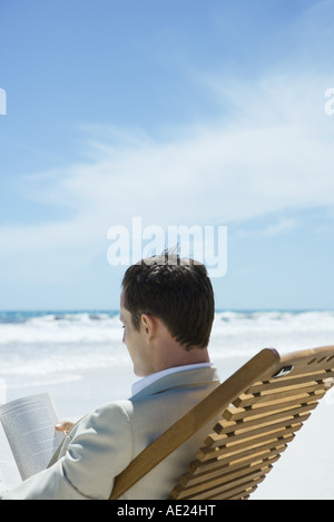 Imprenditore seduto nella sedia a sdraio in spiaggia, lettura, vista posteriore Foto Stock