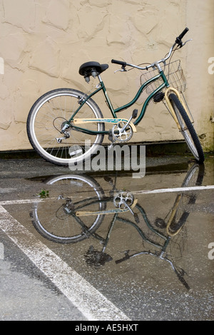 Bicicletta con cestello anteriore in appoggio contro la parete riflettendo contro la pozza nel parcheggio dopo la pioggia ha interrotto Foto Stock