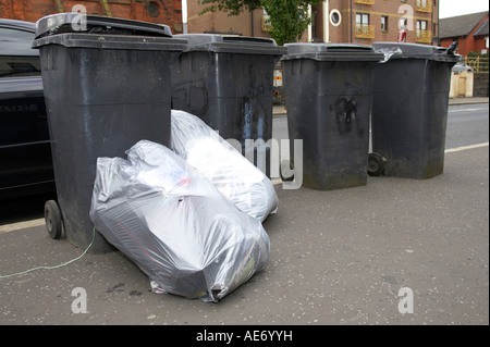 Sacchetti di plastica piena di spazzatura di overflow che giace accanto al grigio scomparti wheelie schierate sul sentiero dalla strada per la raccolta di rifiuti Foto Stock