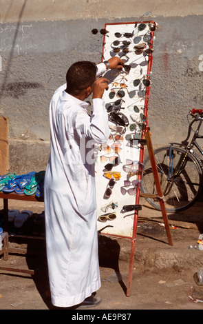 Uomo arabo in un Jelabiya impostazione di un paio di occhiali da sole in stallo il Bazaar Principale, Aswan, Egitto Foto Stock