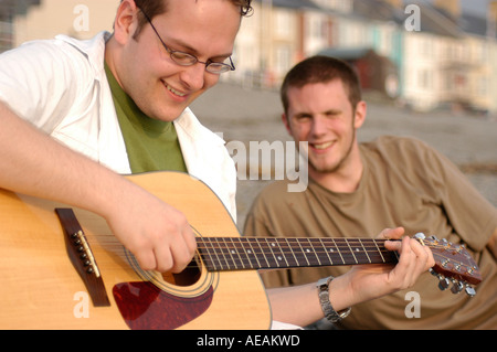 Giovani studenti di sesso maschile a Aberystwyth University suonare la chitarra acustica sulla spiaggia la sera d'estate REGNO UNITO Foto Stock