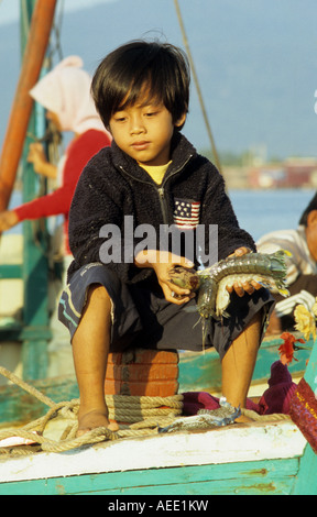 Giovani Cham ragazzo seduto su una barca da pesca tenendo a base di frutti di mare, sul fiume Sanke, Kampot, Cambogia Foto Stock