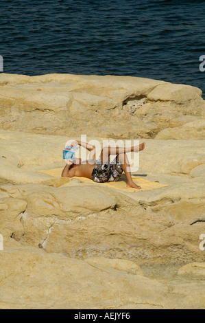 Un uomo a prendere il sole e leggere un libro in costa rocciosa di Sliema città situata sulla costa nord-orientale di Malta nel nord del Distretto di Porto Foto Stock