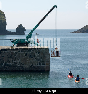 Mullion Due canoisti di lasciare il porto di entrata a parete con workman utilizzando piccole gru per tenere la base di lavoro effettuare riparazioni dopo la tempesta Cornwall Regno Unito Foto Stock