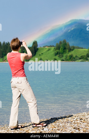 Giovane donna con fotocamera digitale garantisce immagini di spettacolare rainbow su Forggensee, Baviera, Germania Foto Stock