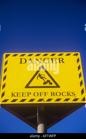 Il giallo e il nero segno contro chiaro profondo cielo blu indicante pericolo tenere Rocks off con simbolo iconico di uomo lo scivolamento sulle rocce Foto Stock