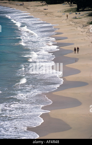 Le piccole onde che si infrangono come navigare sulla curva di spiaggia di sabbia con coppia a piedi nella metà distanza costa sud Caraibi Antigua Foto Stock