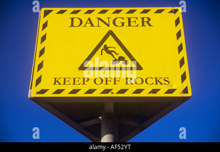 Il giallo e il nero segno contro chiaro profondo cielo blu indicante pericolo tenere Rocks off con simbolo iconico di uomo lo scivolamento sulle rocce Foto Stock