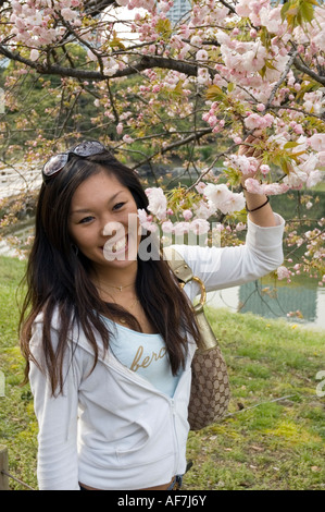 Giovane donna giapponese in piedi vicino a un ramo di fiori di ciliegio in giardino Hamarikyu, Chuo-ku, Tokyo, Giappone Foto Stock