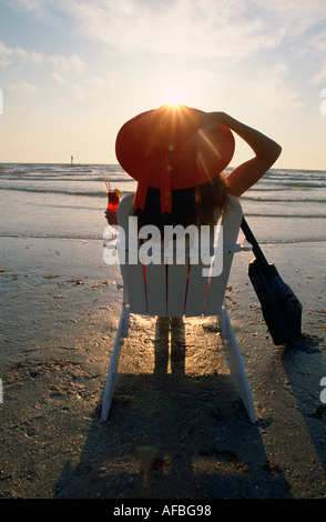 Bruna giovane donna guardando il tramonto su una sdraio sulla spiaggia tenendo un drink congelati e una chitarra elettrica sulla sua destra Foto Stock