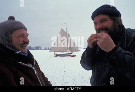 Paesi Bassi Monnickendam uomo sigaretta di rotolamento sul lago ghiacciato Gouwzee con ghiaccio barca a vela in background. Foto Stock