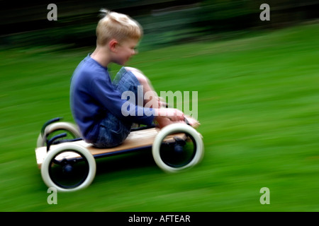 6 anno vecchio ragazzo in sella a una casa fatta buggy o go-kart sulla velocità di erba Foto Stock