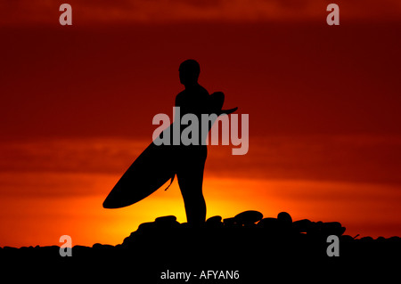 Un surfista passeggiate verso la spiaggia con la sua tavola da surf contro un rosso arancione cielo a sunrise, Devon Foto Stock