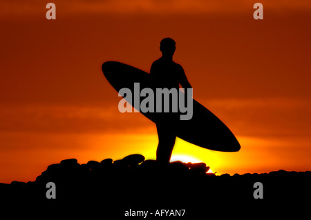 Un surfista passeggiate verso la spiaggia con la sua tavola da surf contro un rosso arancione cielo a sunrise, Devon Foto Stock