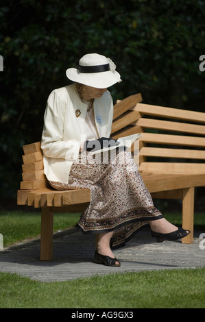 Elegantemente vestito donna di mezza età, indossando guanti e cappello, seduta su una panchina in un parco, lettura. Foto Stock