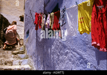 Una donna sale le scale a Chefchaouen, Marocco, con abiti colorati appesi su una linea contro le iconiche pareti dipinte di blu della Medina. Foto Stock