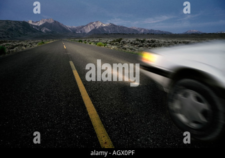 Un auto è sfocata lungo un scurire autostrada durante la guida notturna in una valle vicino alle montagne Foto Stock