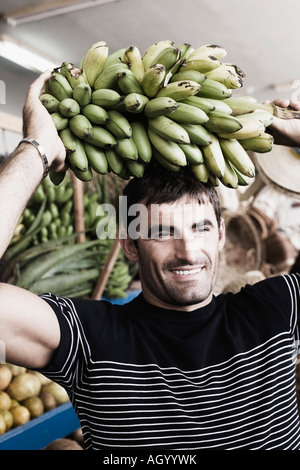 Close-up di una metà uomo adulto che porta un grappolo di banane sul suo capo Foto Stock