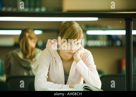 Giovane donna studiare in biblioteca universitaria, tenendo la testa, guardando lontano Foto Stock