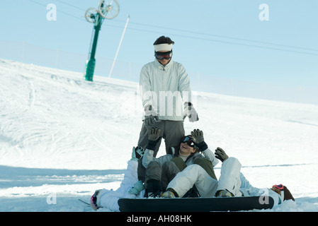 I giovani snowboarders giacente sul terreno raggiungendo per il giovane uomo in piedi dietro di loro Foto Stock