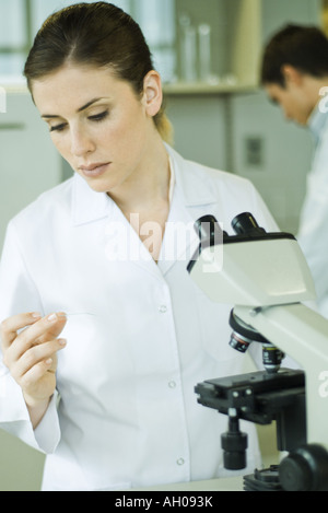 Laboratorio femmina lavoratore in piedi accanto al microscopio, tenendo fino e ispezione della slitta Foto Stock