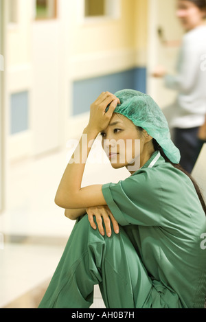 Medical lavoratore seduto sul pavimento nel corridoio di ospedale, tenendo la testa, guardando lontano Foto Stock