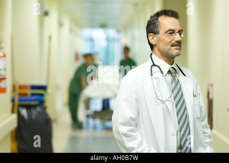 Medico di sesso maschile che guarda lontano, sorridente, ospedale corridoio in background Foto Stock