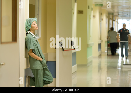 Infermiera femmina in ospedale, appoggiata contro il muro con le mani nelle tasche, vista laterale Foto Stock