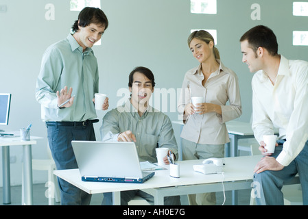 Quattro soci di affari in ufficio, guardando il computer portatile, tenendo tazze da caffè Foto Stock