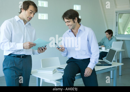 Uomini d'affari in ufficio, uno tramite telefono cellulare, sorridente Foto Stock