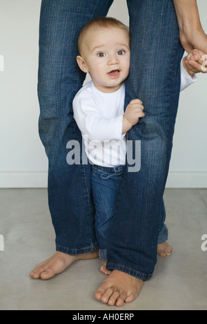 Il bambino in piedi fra una società madre per le gambe, guardando la fotocamera Foto Stock