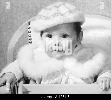 Vecchia famiglia vintage fotografia istantanea della bambina indossa pelliccia cappotto rifilato e lana cofano seduto nella sedia alta Foto Stock