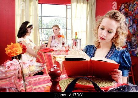 Giovane donna leggendo un libro con due giovani donne di pranzare in un ristorante dietro di lei Foto Stock