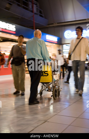 Un membro del personale dell'aeroporto spinge una sedia a rotelle del passeggero di partenza attraverso lounge area dello shopping a Londra Heathrow Terminal 4 Foto Stock