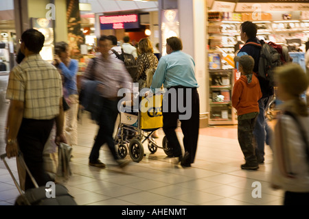 Un membro del personale dell'aeroporto spinge una sedia a rotelle del passeggero di partenza attraverso lounge area dello shopping a Londra Heathrow Terminal 4 Foto Stock