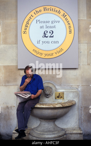 Un uomo fuma una sigaretta e legge un giornale al di sotto del fondo di un segno di sollevamento al di fuori del British Museum di Londra, Regno Unito. Foto Stock