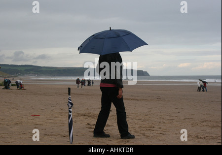 Spiaggia di sabbia sotto la pioggia, sta un uomo sotto un ombrello che nasconde la sua faccia. Vi è un altro ombrellone, arrotolato, accanto a lui Foto Stock