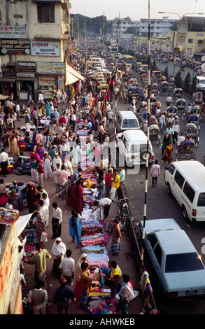 Traffico serale in una strada trafficata con mercato dell'abbigliamento sul marciapiede a Dhaka, Bangladesh Foto Stock