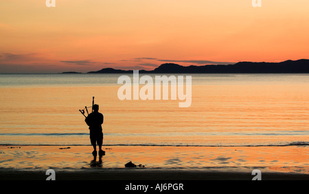 Un bagpiper stagliano contro il tramonto sulla spiaggia Arisaig nelle highlands con Skye oltre Foto Stock