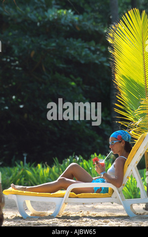 Giovane donna sdraiata su bedchair sulla spiaggia, indossa foulard blu e occhiali da sole Foto Stock