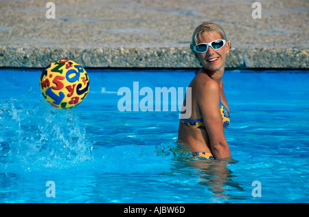 Giovane donna che gioca con la palla in piscina, indossando bikini colorati e occhiali da sole Foto Stock