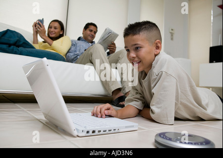 Boy utilizzando laptop sul pavimento nel soggiorno, madre e padre sul divano, vista terra Foto Stock